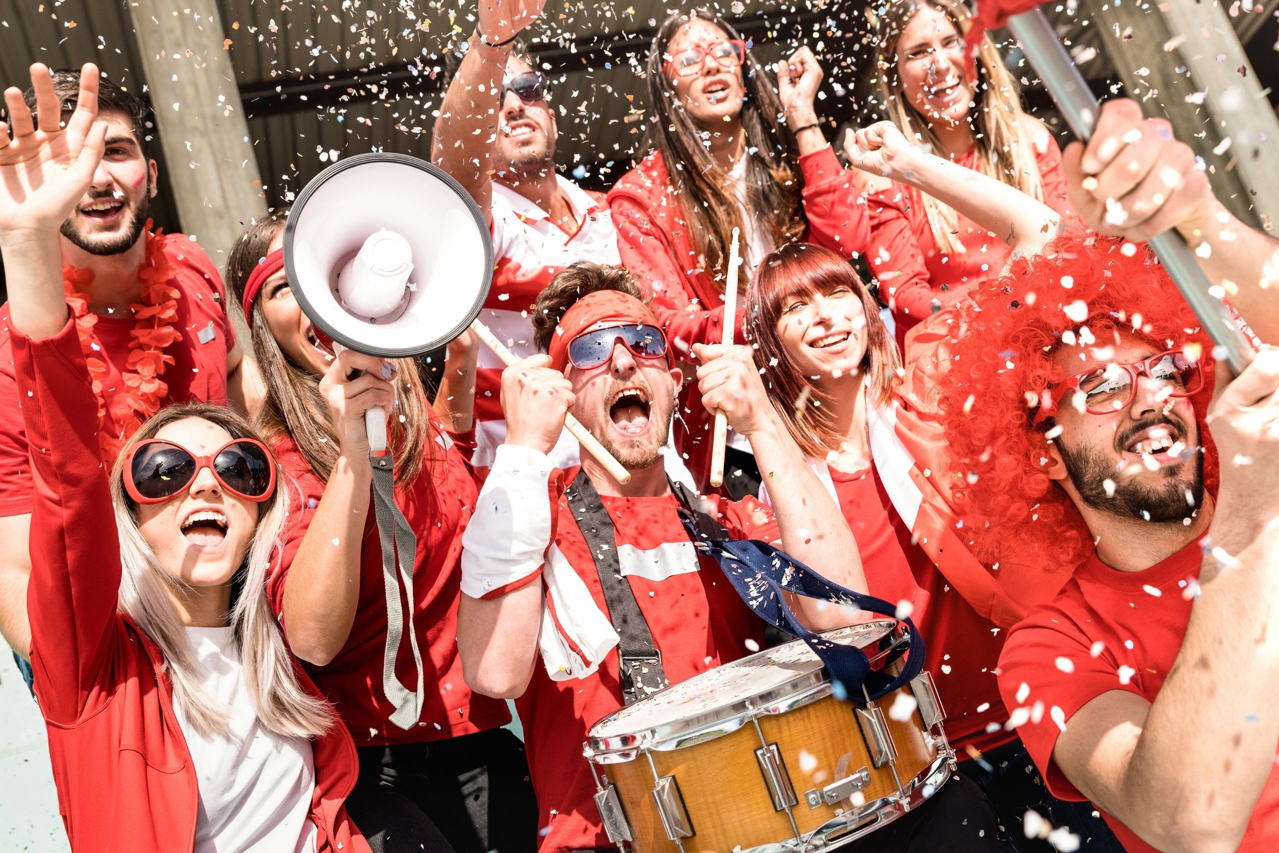 Group of enthusiastic people dressed in red and white, cheering and celebrating with confetti, a megaphone, and a drum - capturing the energy, unity, and excitement of collective support at a rally or event.